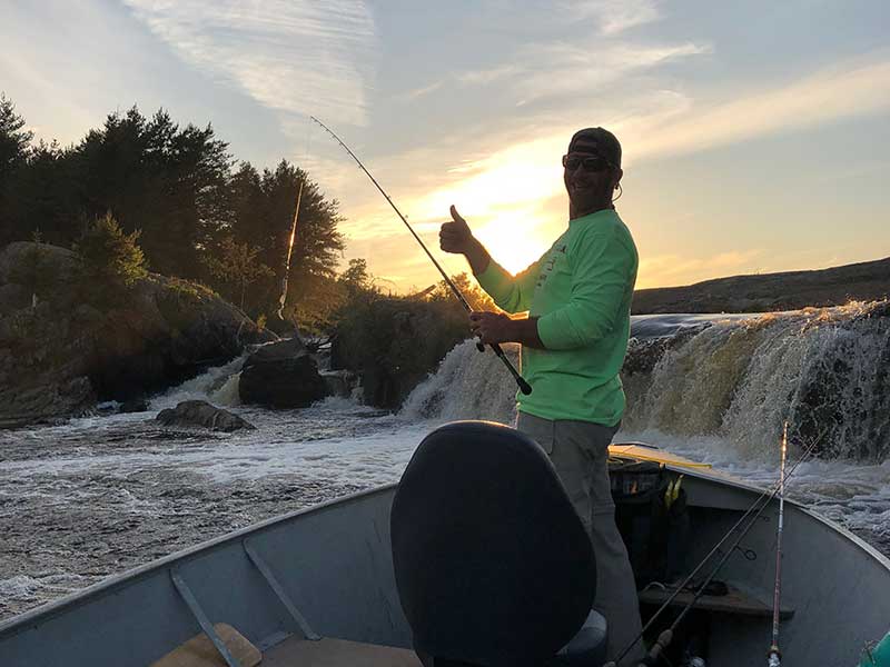 Fisherman giving thumbs up from his boat in front of a waterfall at sunset.