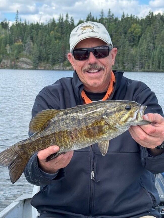 Man wearing sunglasses while holding a fish in the front of a boat and he has a Halley's Camps hat on.