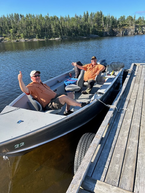 2 men in a boat waving and thumbs up for the picture on a sunny day at Halley's Camps and the boat is tied to the dock