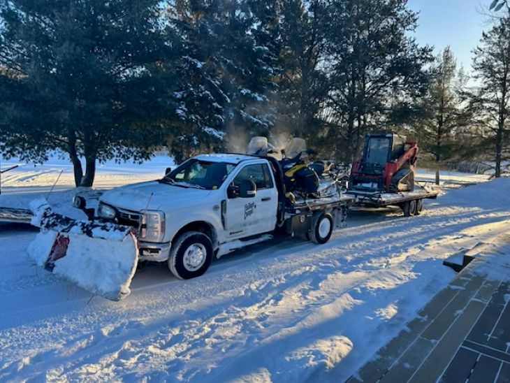 The 1 ton warming up on a cold day with the 2 sleds on the flat deck and the skid steer on the flat deck with blue skies and snow on the ground and trees in the background