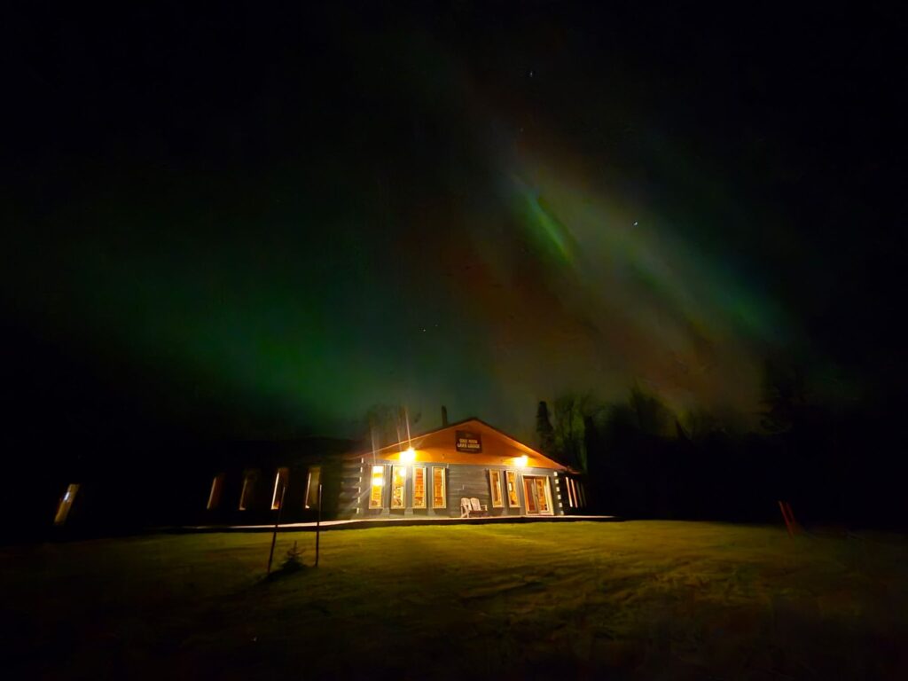 Picture of One Man Lake Lodge at night and you can see the grass in the front year with the lights on the outside and the northern lights are green, blue, and pink in the sky behind the lodge