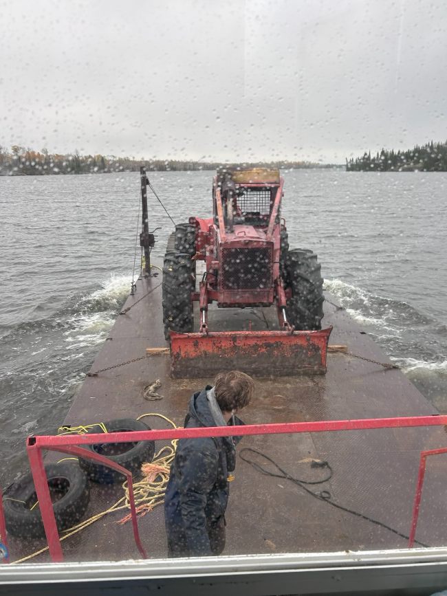 Flat barge with a skidder on it on a cloudy day with a worker walking and checking on things while barge is in the middle of the lake
