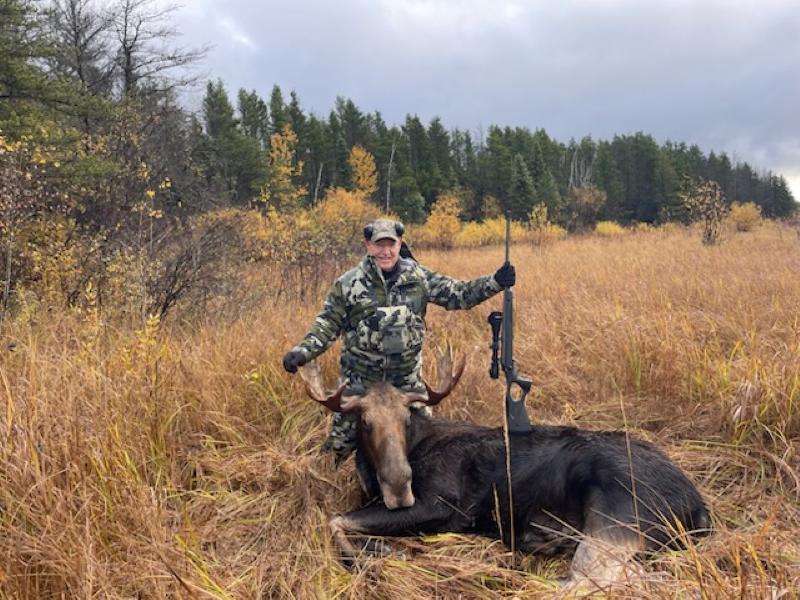 Man standing behind his bull moose with one hand on the antler and the other holding on to his rifle that is propped on the moose's back
