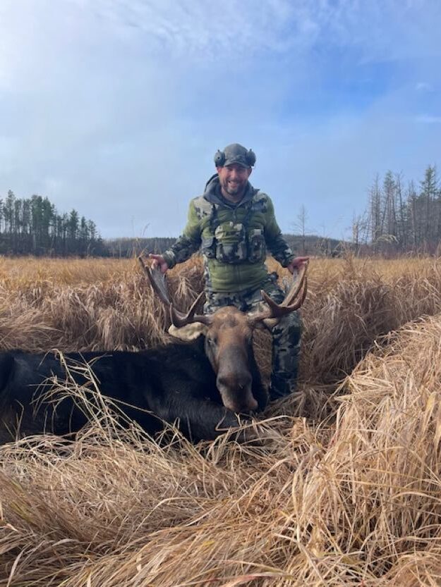 Man standing in a swamp of brown cat tails (weeds) on a blue sunny day while he holds onto his moose antlers