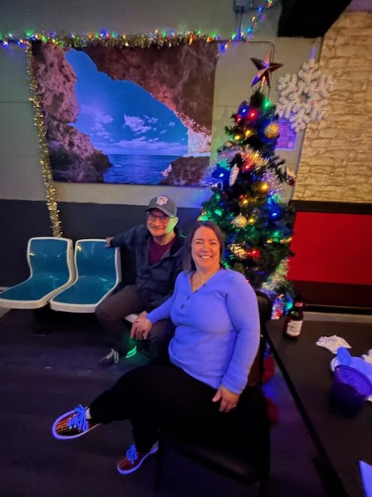 Husband and wife sitting at a bowling alley smiling for the picture with a Christmas tree in the background