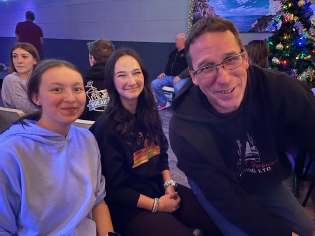 2 girls and a man smiling for a photo in a bowling alley with people behind them