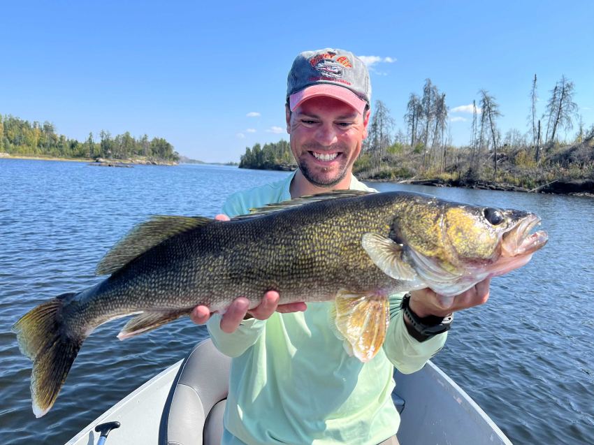 Man with big smile holding his walleye at the front of the boat while fishing at Halley's Camps. The lake is blue and so is the sky and the fish is glimmering with yellows and greens from the sun.