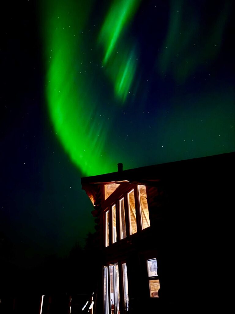 Halley's Trapline Lake Outpost cabin at night with green northern lights in the skies with stars while inside the cabin is lit up.