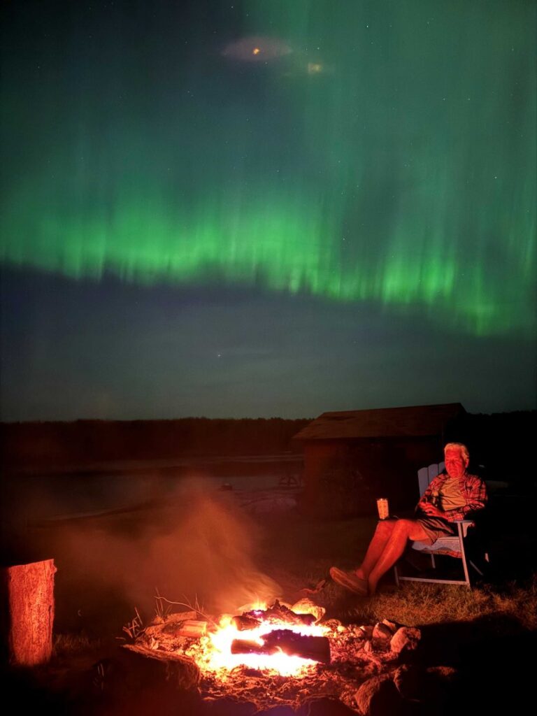 Man sitting next to a fire with the lake behind him with green northern lights in the skies.